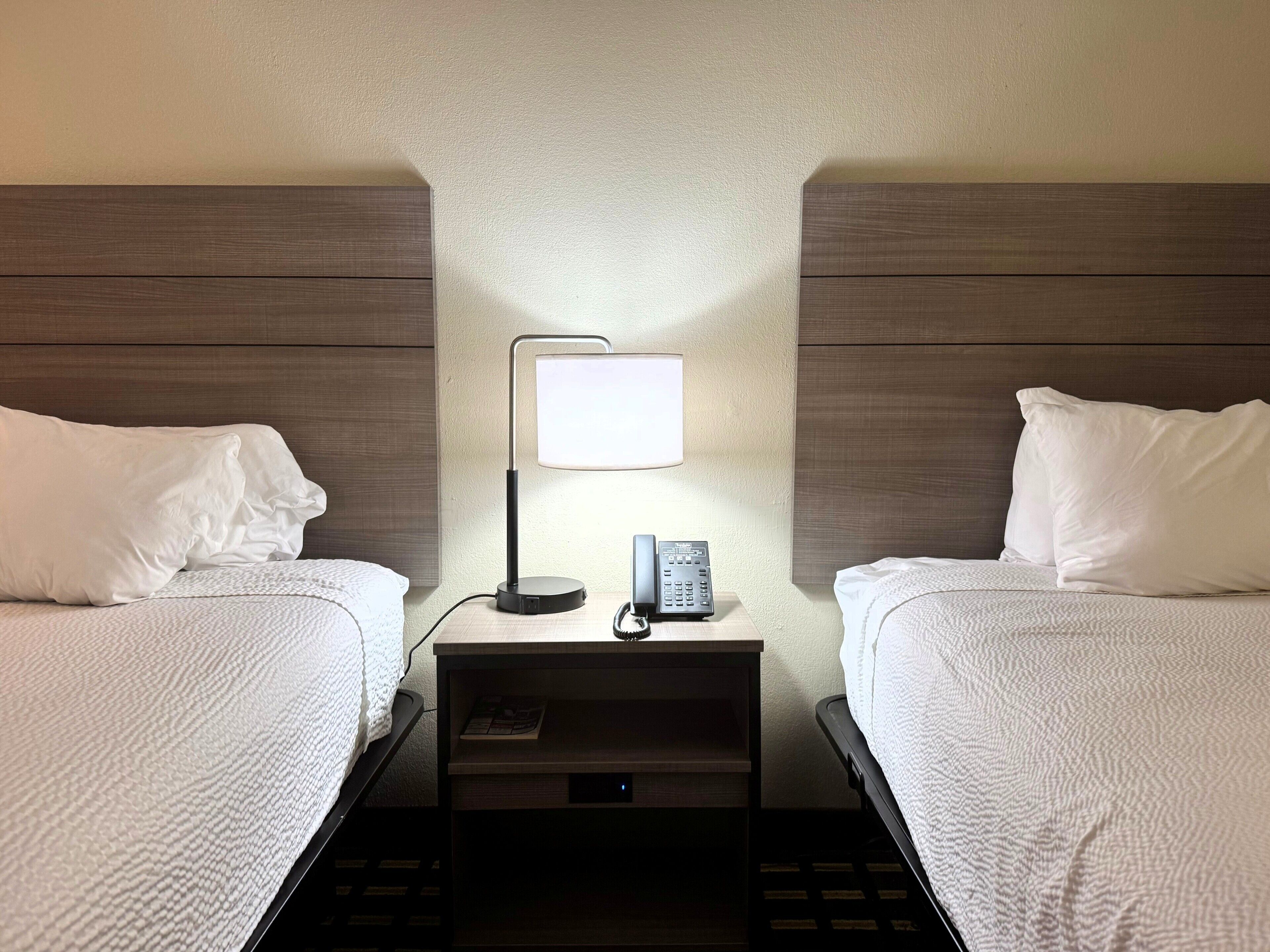 A hotel room with two beds, a nightstand with a lamp, a phone, and a small drawer.