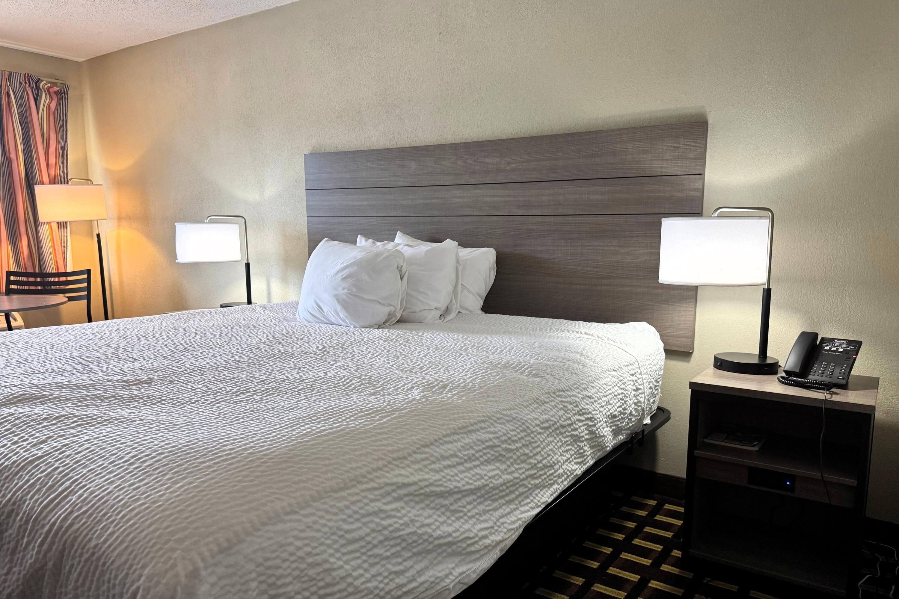 A hotel room with a large bed, two bedside lamps, a bedside phone, and a small table with a chair.