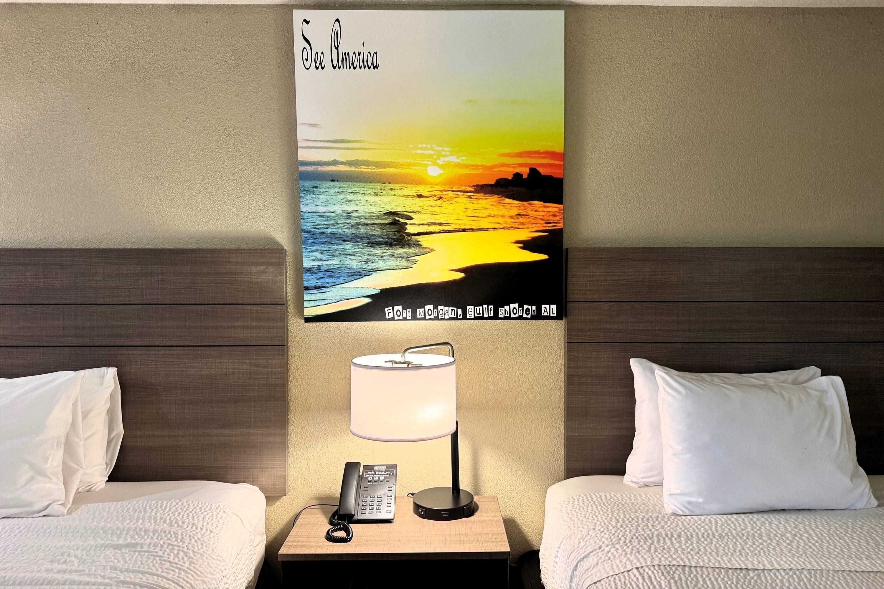 Hotellihuone, jossa on sänky, yöpöytä lampun kanssa, puhelin ja auringonlaskun kuva seinällä.