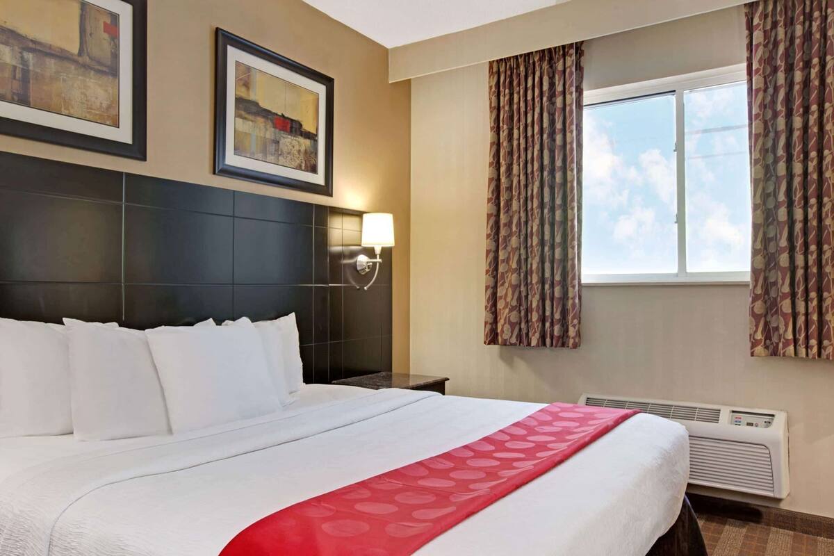 A hotel room with a large bed, a headboard, a wall-mounted lamp, a window with curtains, and a painting on the wall.
