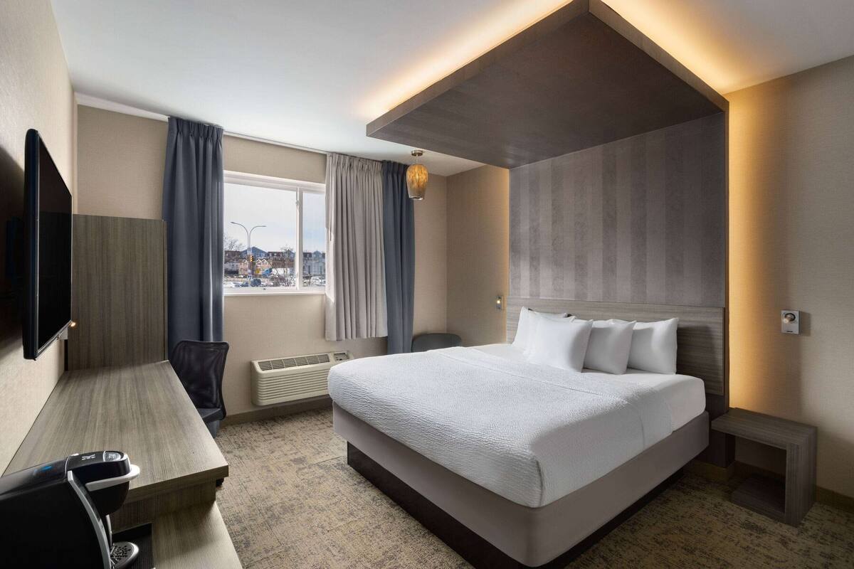 A modern hotel room with a large bed, a desk, a flat-screen TV, and a view of the city.