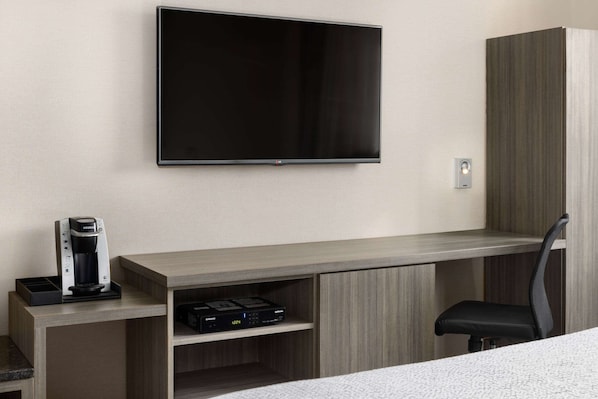 A hotel room with a flat-screen TV mounted on the wall, a coffee maker, a Samsung sound system, and a black office chair.