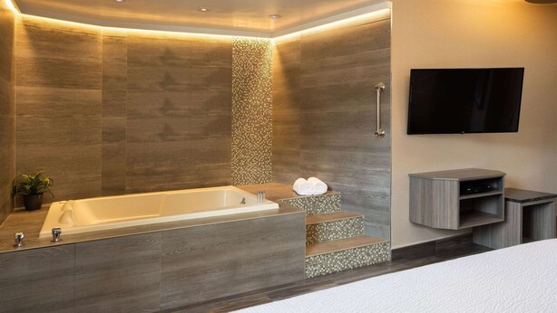Un baño moderno con una bañera grande, una zona de ducha con un escalón, un televisor de pantalla plana montado en la pared y un tocador minimalista con espejo.