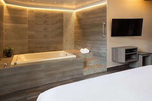 Un baño moderno con una bañera grande, una zona de ducha con un escalón, un televisor de pantalla plana montado en la pared y un tocador minimalista con espejo.