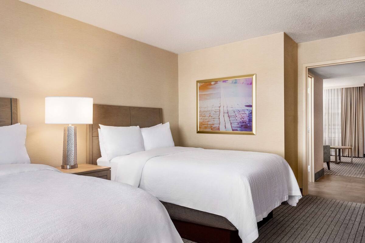 A hotel room with two beds, a nightstand with a lamp, a painting on the wall, and a view of the hallway.