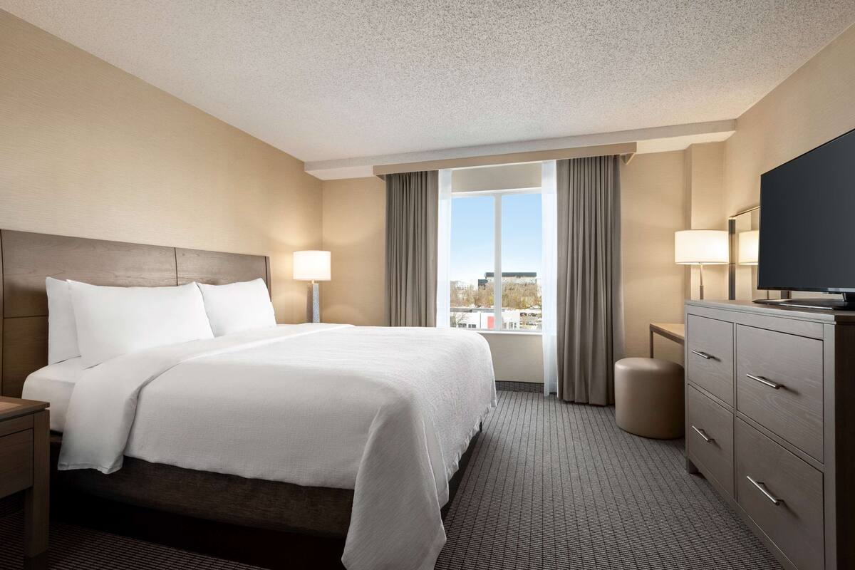 A hotel room with a large bed, a TV, a desk, and a view of the city.