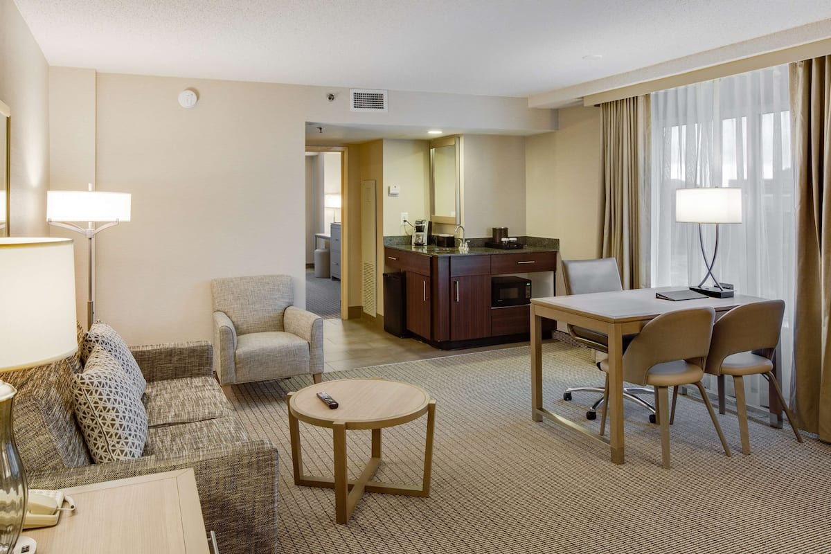 A hotel room with a living area, a dining table with chairs, a kitchen area, and a large window with curtains.