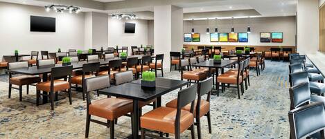A hotel conference room with multiple tables and chairs, a patterned carpet, and a large screen on the wall.
