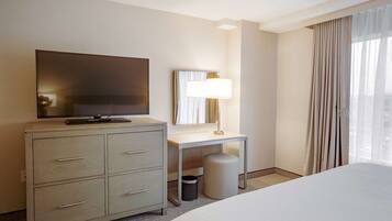 A hotel room with a bed, a TV on a dresser, a desk with a lamp, and a window with curtains.