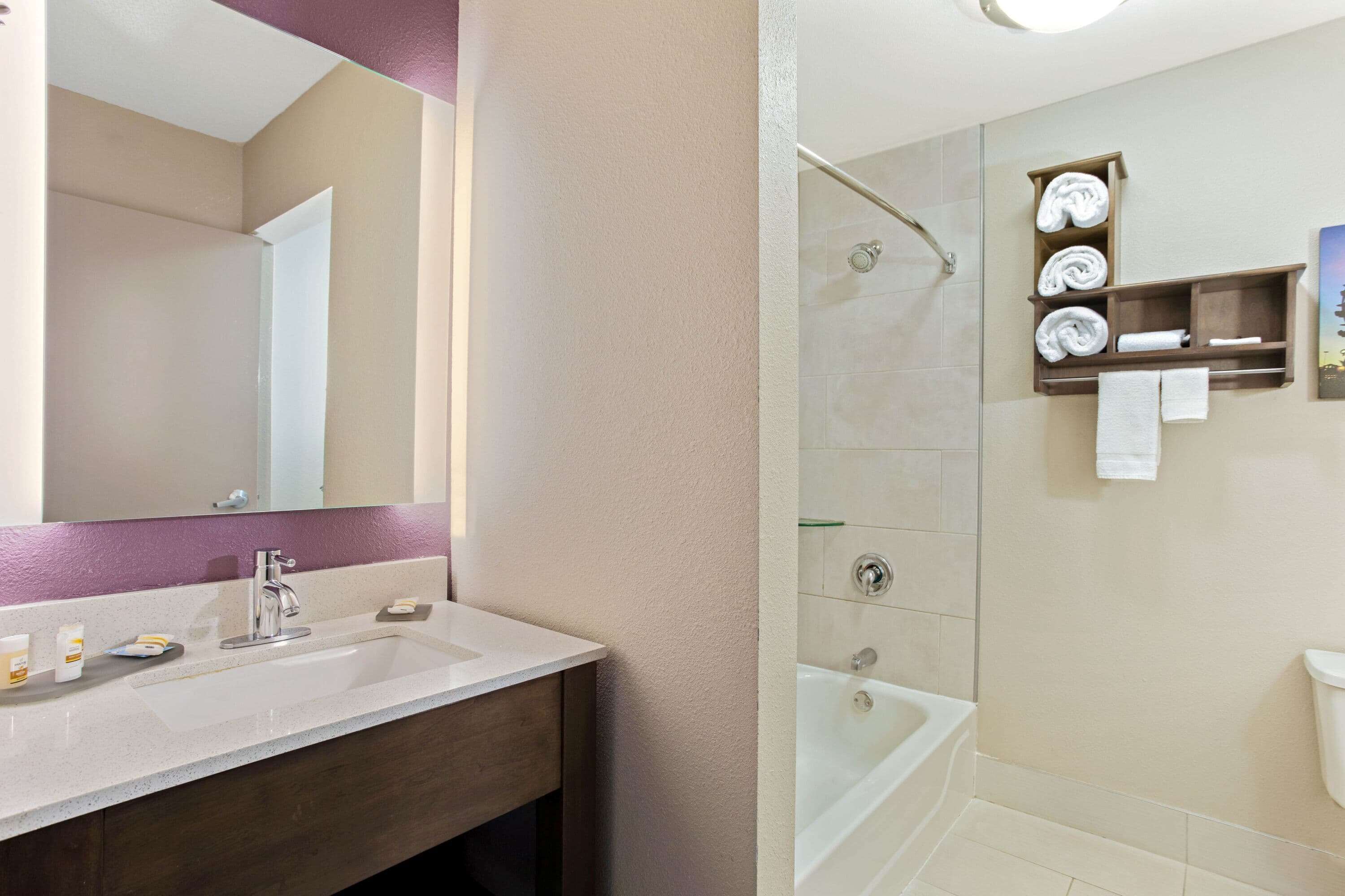 Une salle de bain avec un lavabo blanc, un miroir et un espace douche doté d’une porte en verre. On peut voir des serviettes et des articles de toilette.