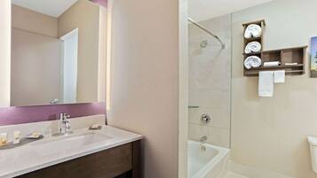 Une salle de bain avec un lavabo blanc, un miroir et un espace douche doté d’une porte en verre. On peut voir des serviettes et des articles de toilette.