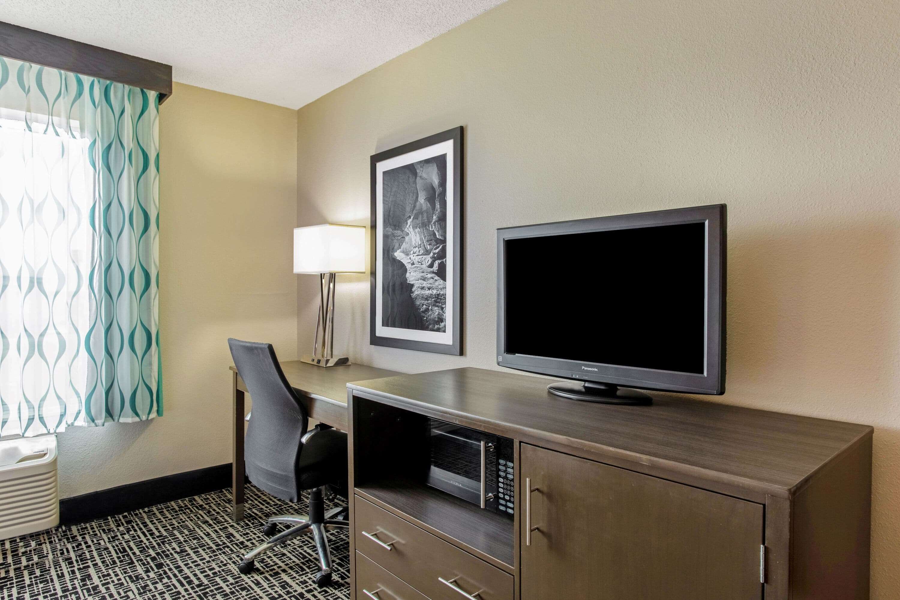 A hotel room with a desk, chair, television, and artwork on the wall.