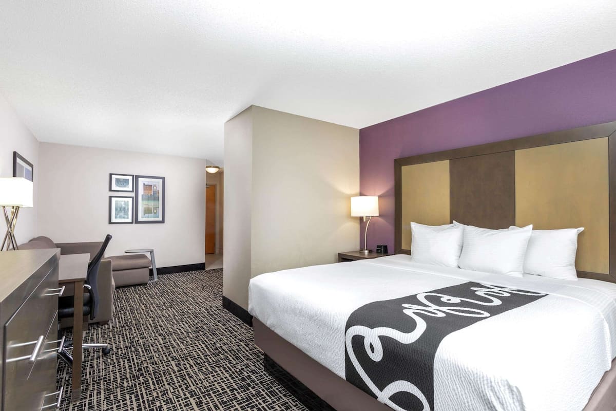 A hotel room with a large bed, a desk with a chair, a lamp, and framed pictures on the wall.