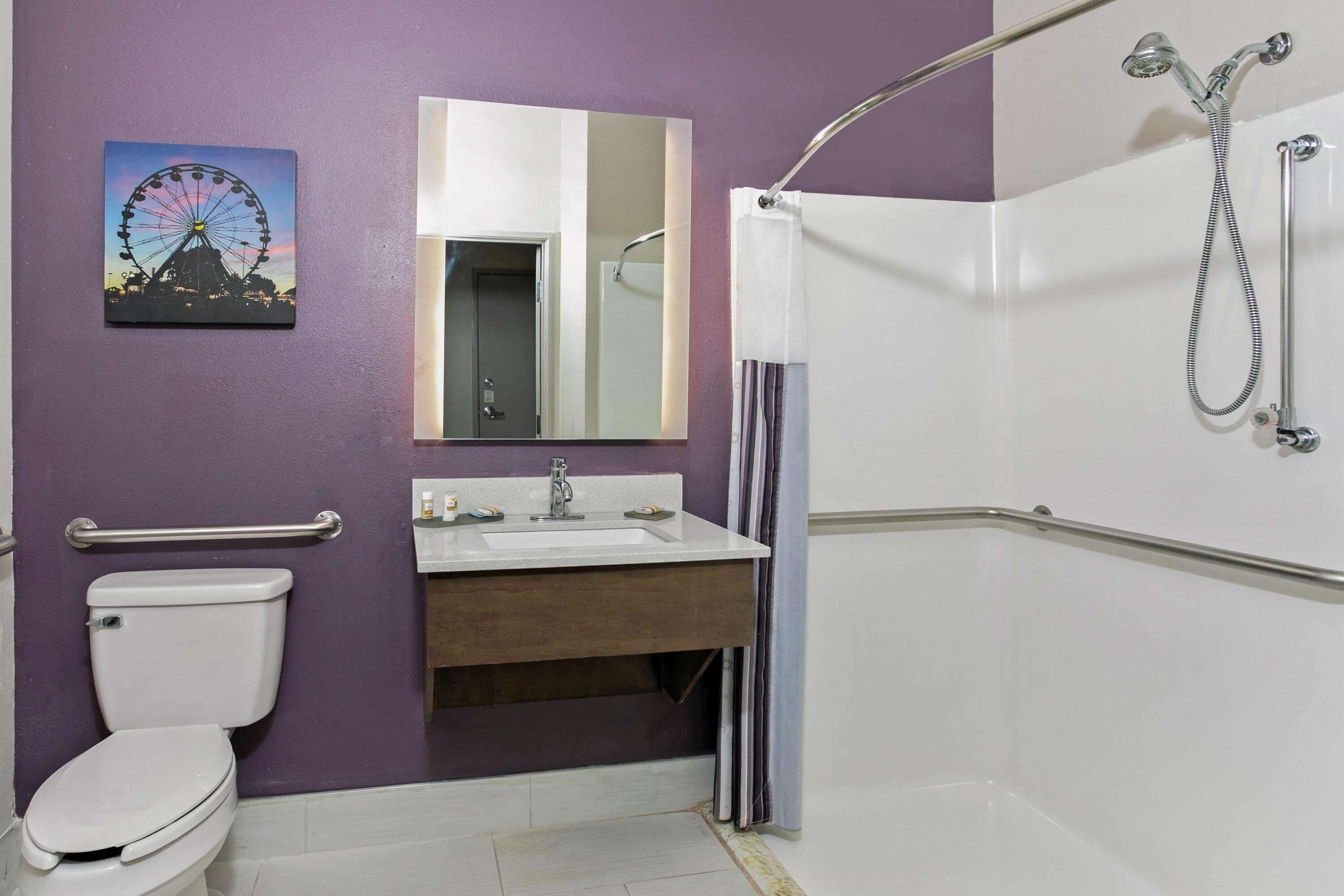 A bathroom with a toilet, sink, mirror, shower, and a wheelchair-accessible grab bar.