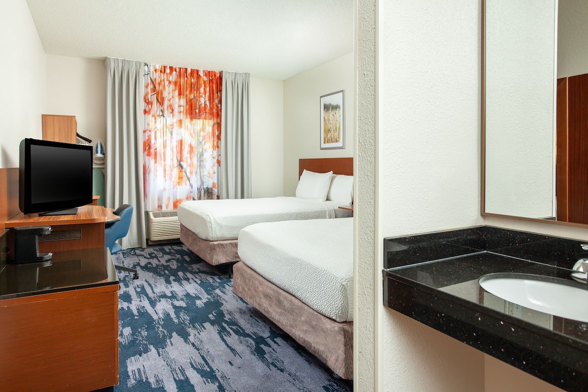 A hotel room with a desk, two beds, a television, a bathroom with a sink and mirror, and a window with curtains.
