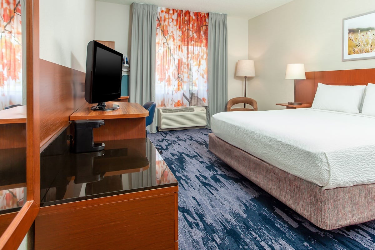 A hotel room with a bed, a desk with a computer monitor, a chair, a lamp, and a window with curtains.