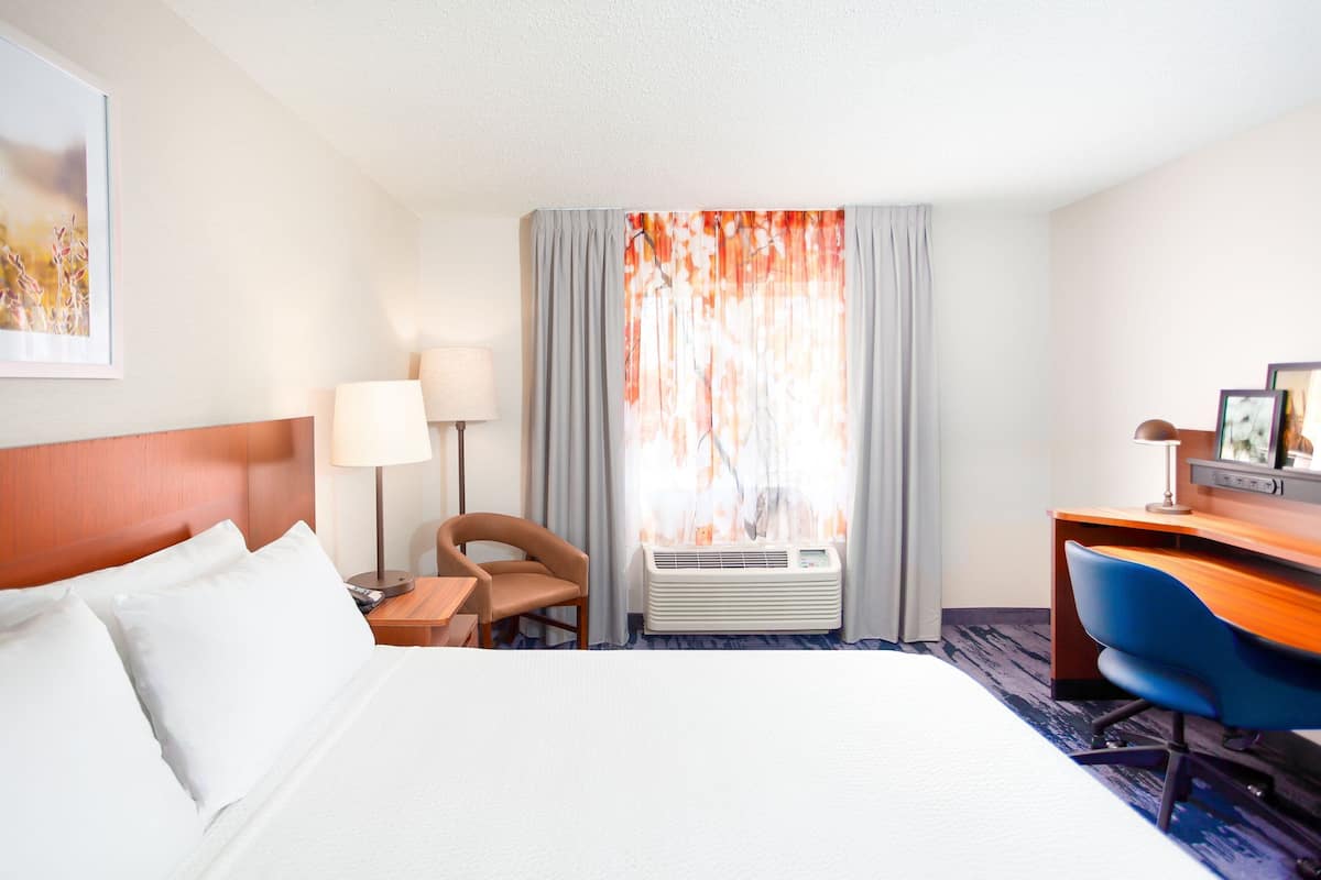 A hotel room with a bed, a desk, a chair, a lamp, and a window with curtains.
