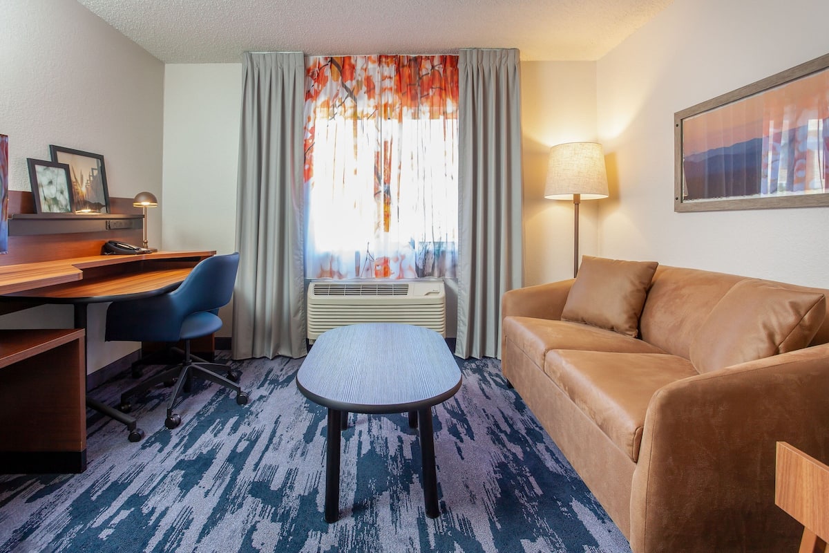A hotel room with a desk, a blue office chair, a brown sofa, a patterned carpet, a window with curtains, an air conditioning unit, and a wall-mounted lamp.