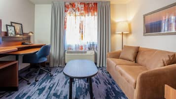 A hotel room with a desk, a blue office chair, a brown sofa, a patterned carpet, a window with curtains, an air conditioning unit, and a wall-mounted lamp.