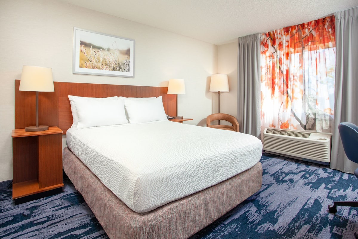 A hotel room with a large bed, two bedside lamps, a headboard, a framed picture on the wall, a window with curtains, and a chair.