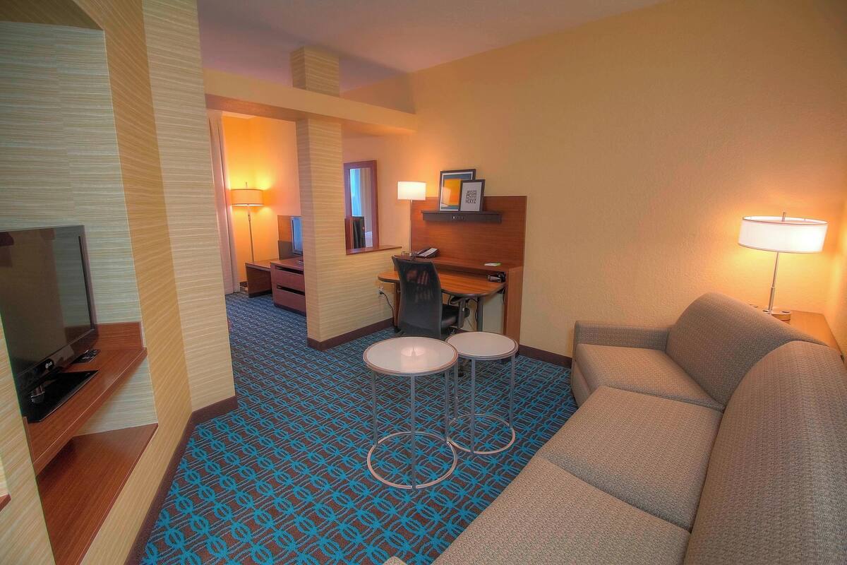 Habitación de hotel con sofá, dos mesitas, escritorio, televisor y alfombra estampada.