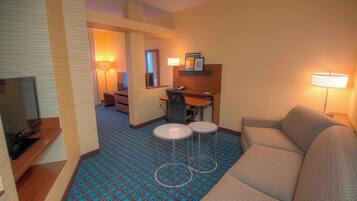 Quarto de hotel com sofá, duas mesinhas, escrivaninha, televisão e carpete estampado.