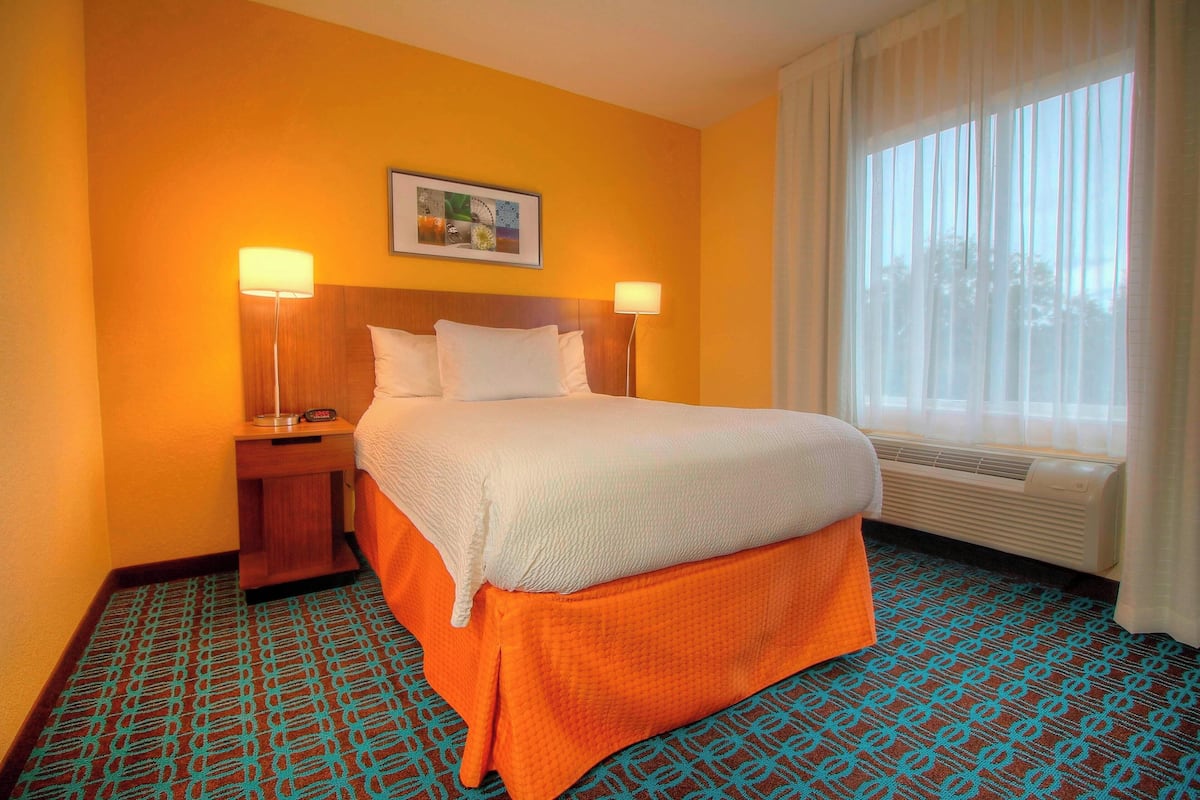 Una habitación de hotel con una cama, dos lámparas de noche, un cuadro enmarcado en la pared, una ventana con cortinas y alfombra estampada.