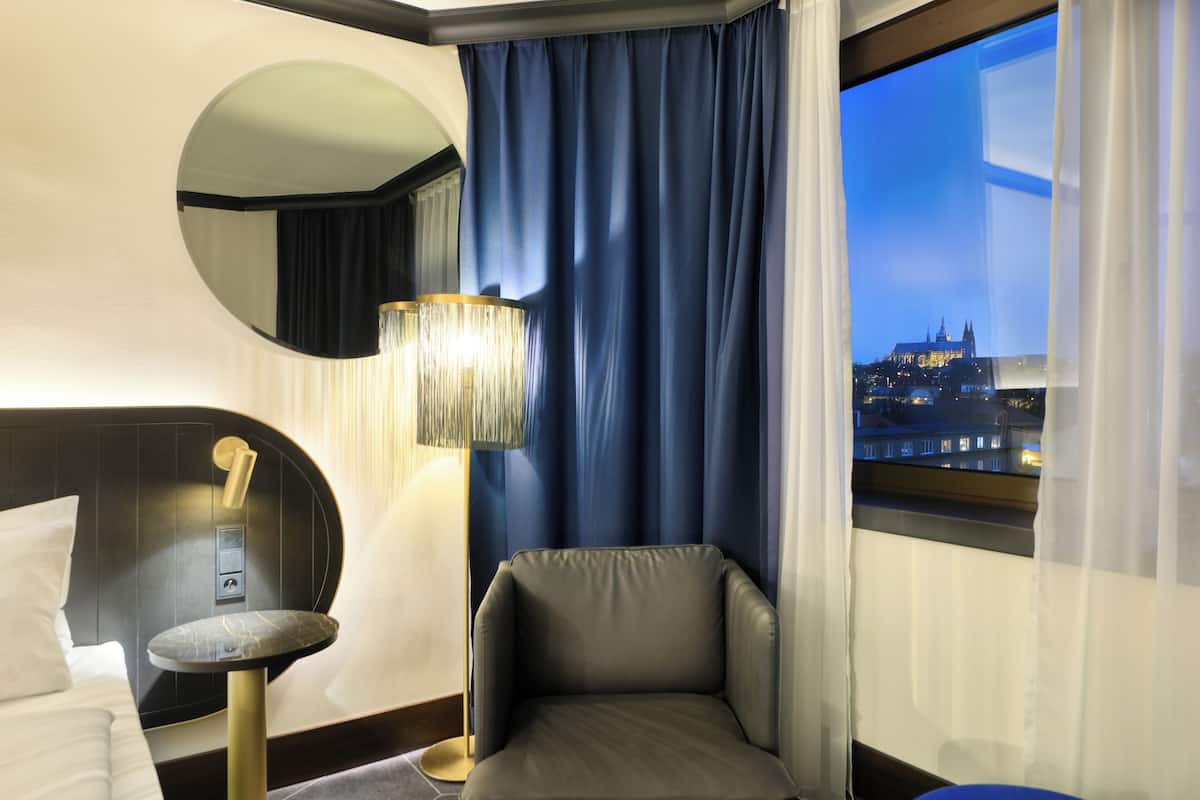 A hotel room with a bed, a round mirror, a small table, a chair, a lamp, and a view of a cityscape at night.