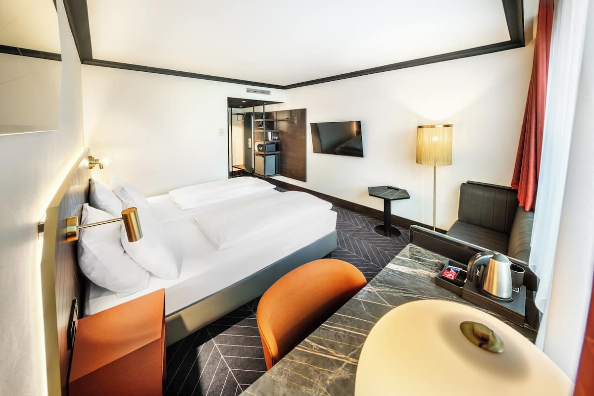 A hotel room with a bed, a desk, a chair, a TV, a lamp, and a bathroom.