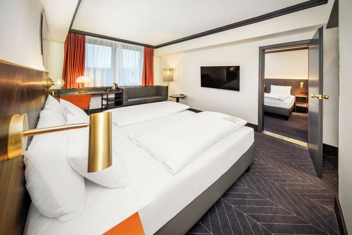 A modern hotel room with a large bed, a desk with a lamp, a TV, and a view of another room.