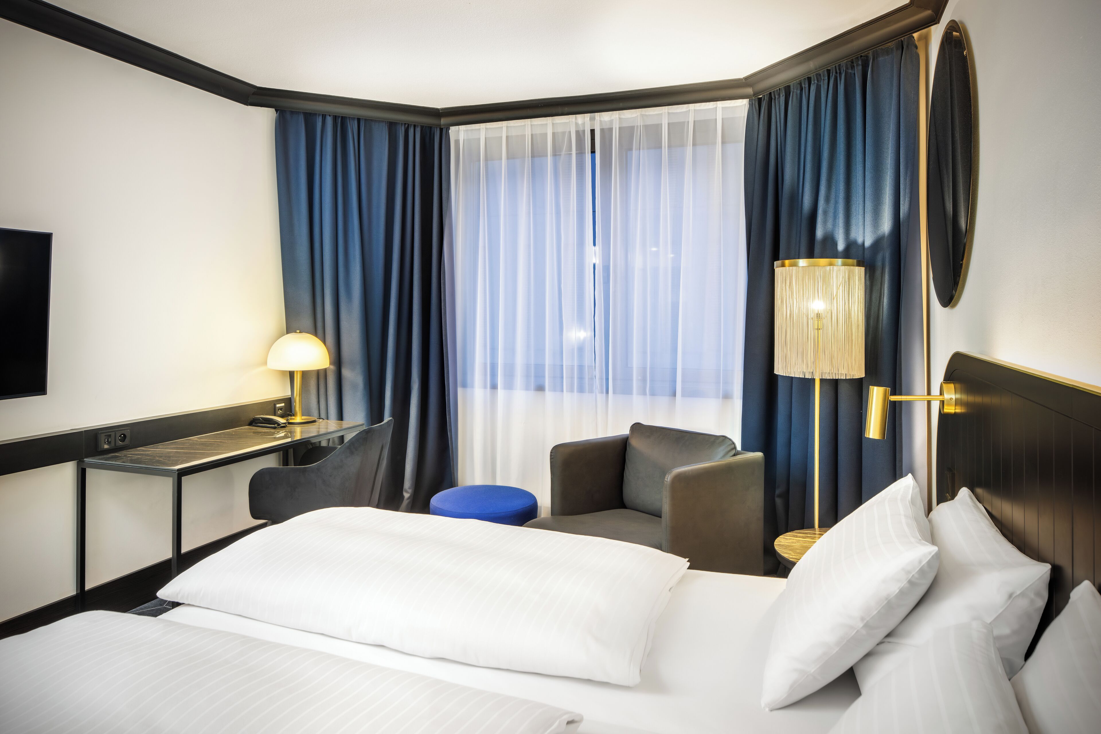 A hotel room with a bed, a desk with a lamp, a chair, and a large window with curtains.