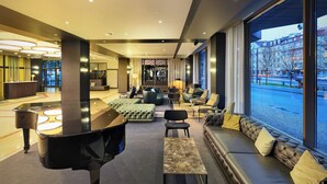 Un elegante vestíbulo de hotel moderno con un piano de cola, asientos cómodos y ventanas grandes que ofrecen vistas a la calle.