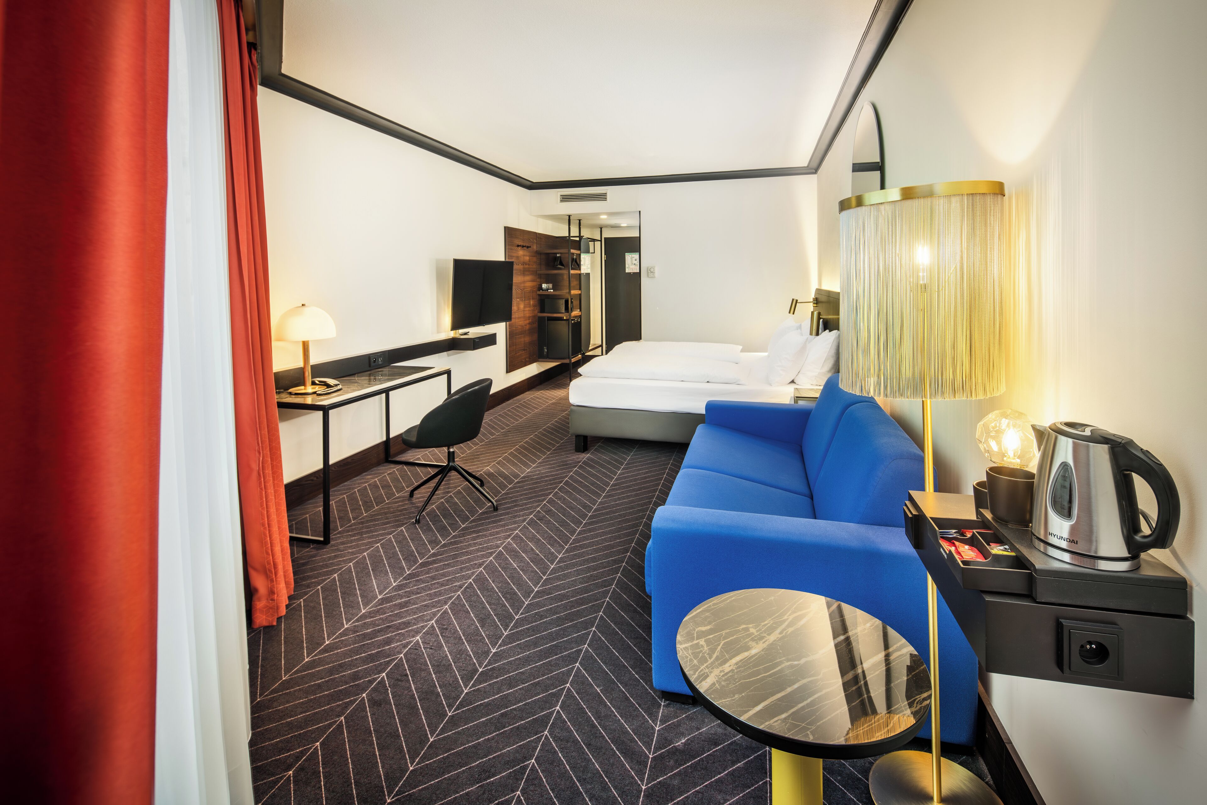 A hotel room with a bed, a blue sofa, a desk with a lamp, a TV, and a coffee maker.