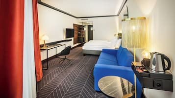 A hotel room with a bed, a blue sofa, a desk with a lamp, a TV, and a coffee maker.