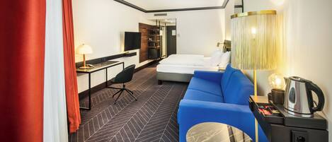 A hotel room with a bed, a blue sofa, a desk with a lamp, a TV, and a coffee maker.
