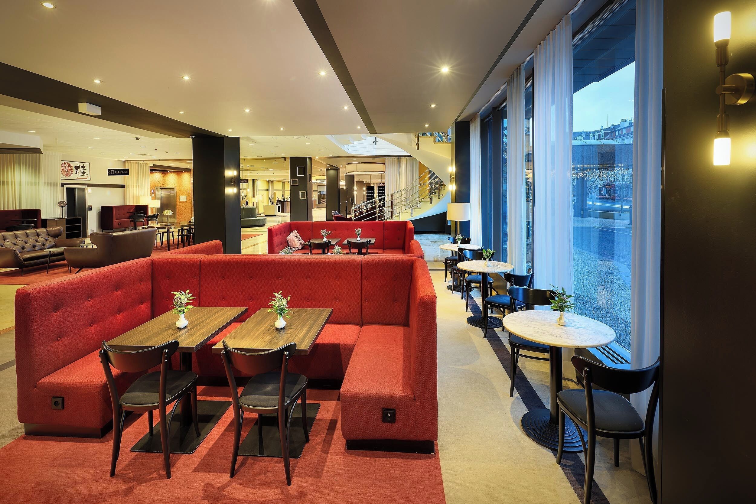 A modern hotel lobby with red seating, tables, and a staircase leading to an upper level.