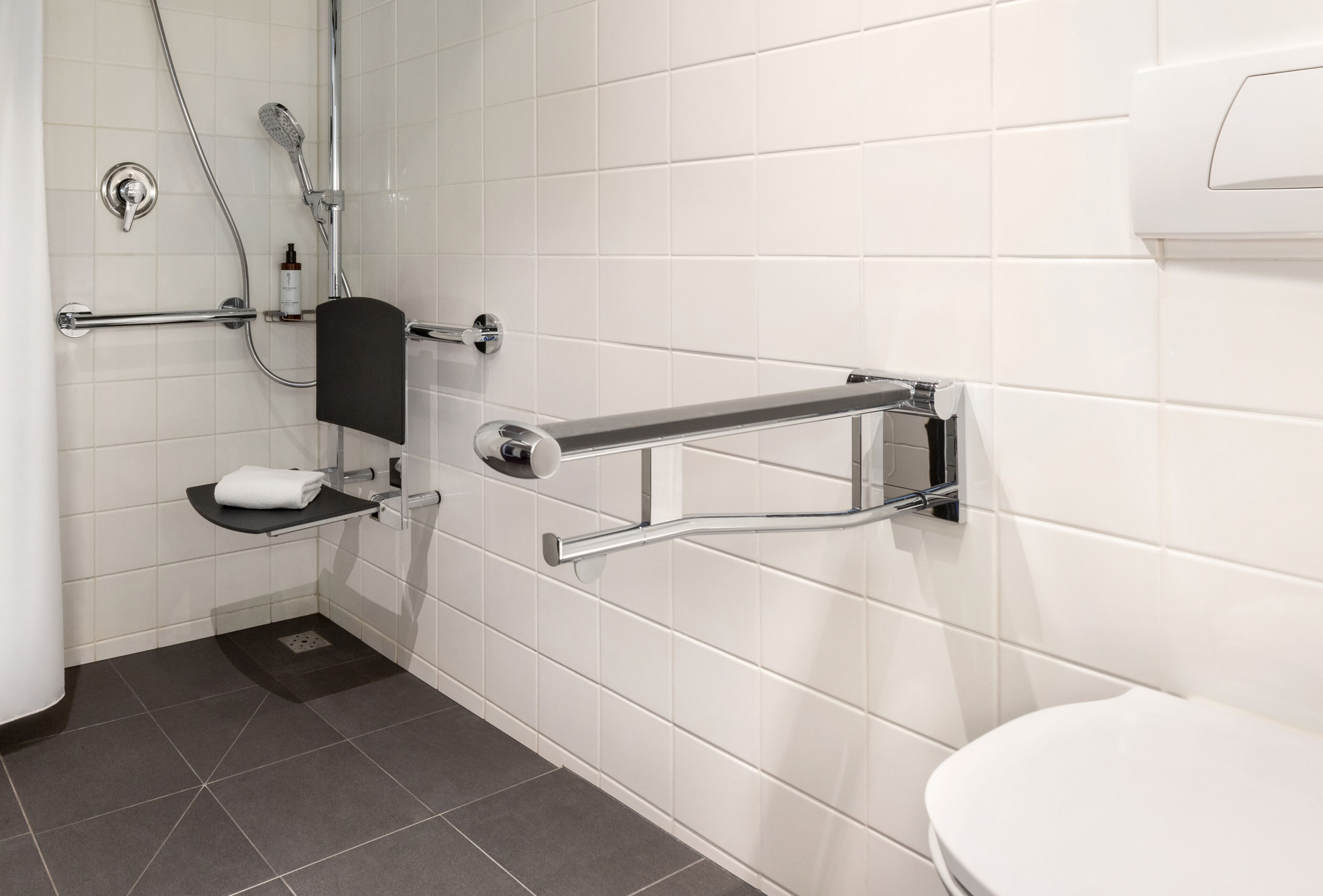 Un baño moderno con paredes de azulejos blancos, inodoro blanco, ducha con cabezal de ducha manual y barra de apoyo cerca del inodoro.