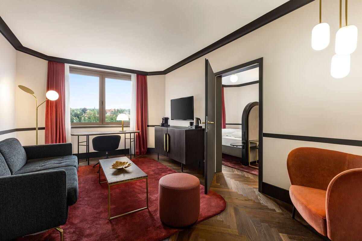 A modern hotel room with a sofa, ottoman, and a desk with a chair. There is a TV, a wardrobe, and a bed with a headboard.