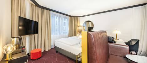 A hotel room with a bed, a red ottoman, a sofa, a TV, a lamp, and a desk with a chair.