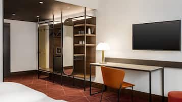 Una habitación de hotel con una cama, un escritorio con silla, un televisor, un armario y un espejo.
