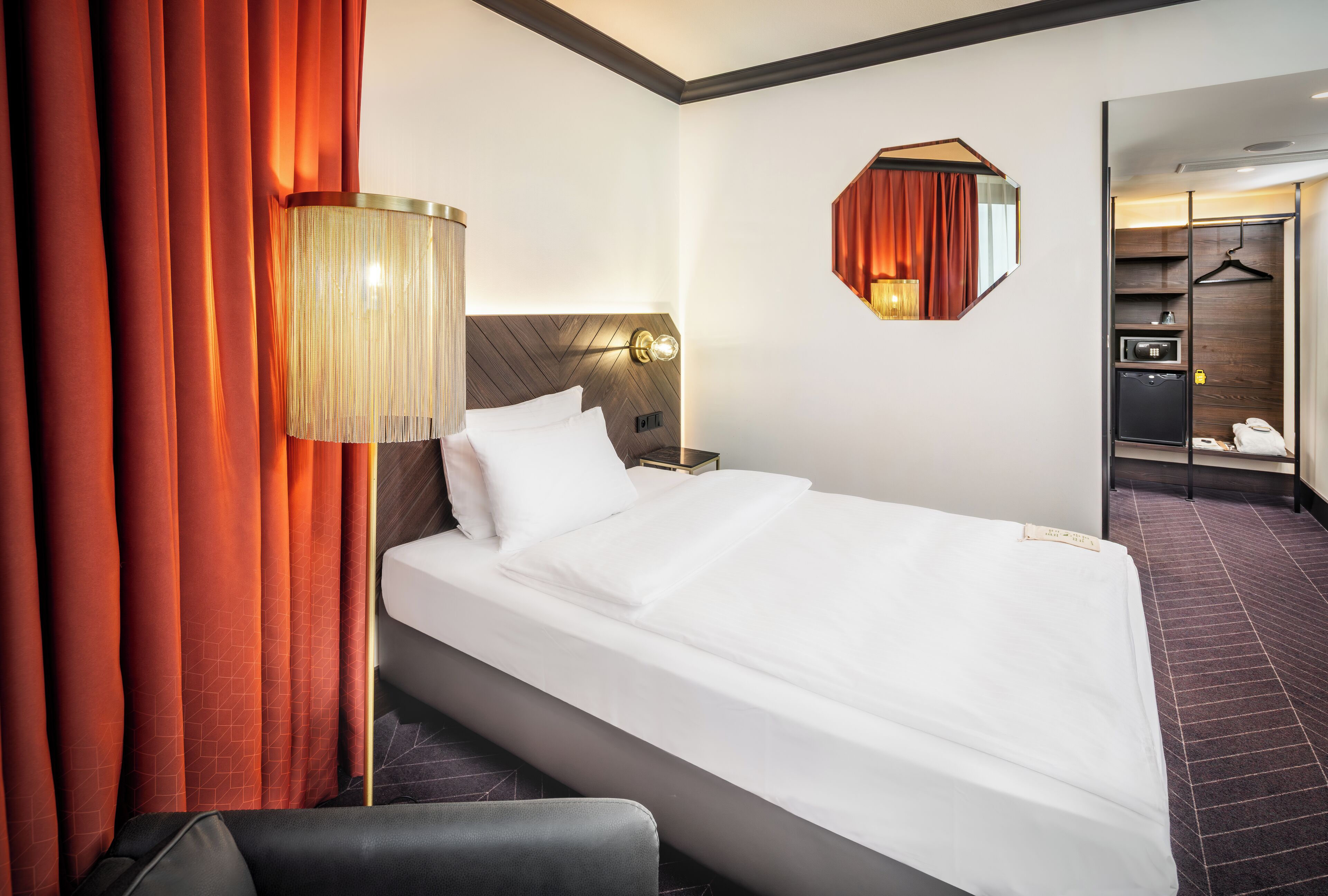Quarto de hotel com cama, mesa de cabeceira, luminária, espelho e guarda-roupa.