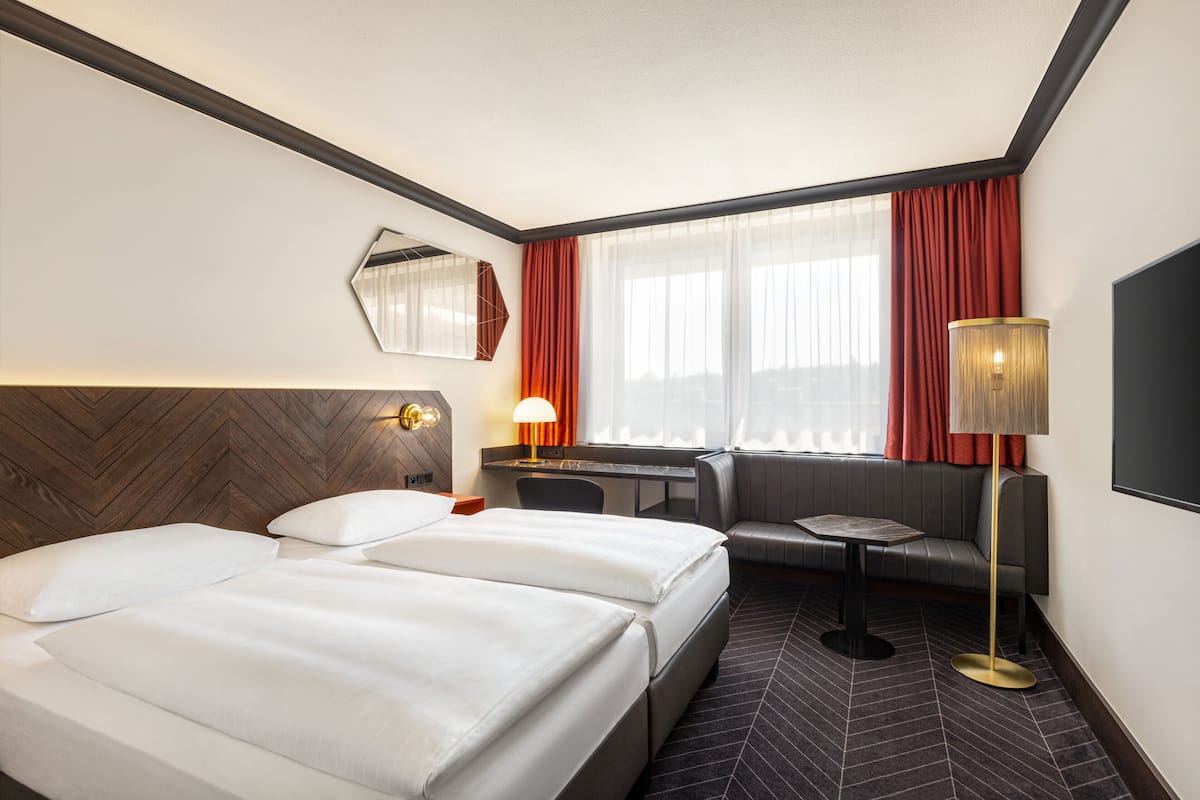 A hotel room with two beds, a desk, a sofa, a lamp, and a large window with curtains.