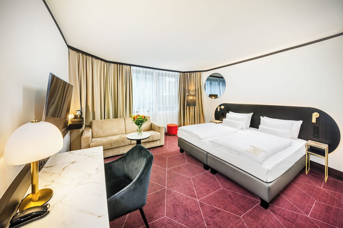 A hotel room with a bed, a desk with a lamp, a chair, a round mirror, and a small table with flowers.
