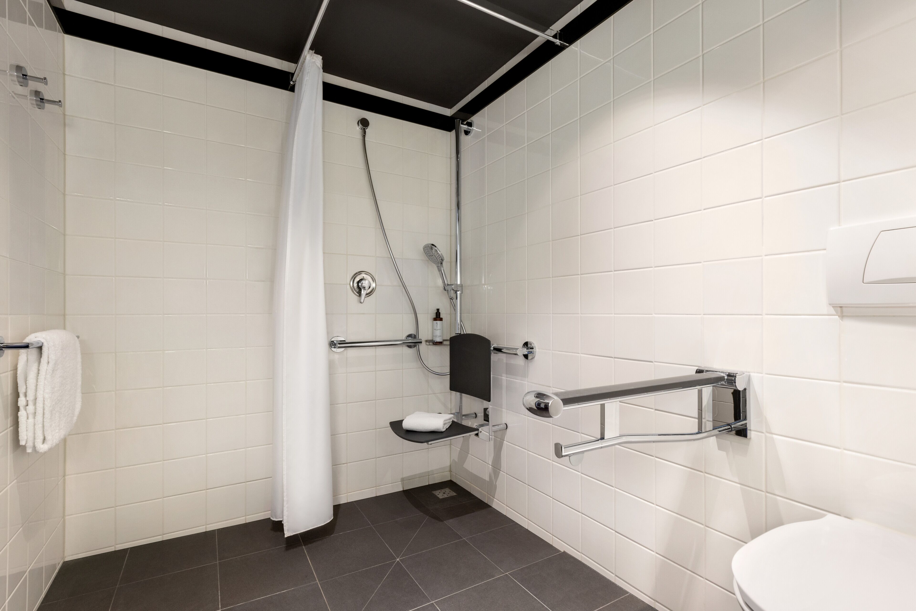 A modern bathroom with white tiles, a shower with a curtain, a grab bar, and a toilet.