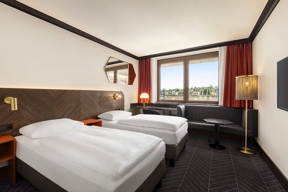 A hotel room with two beds, a desk, a chair, a sofa, a TV, and a window with curtains.
