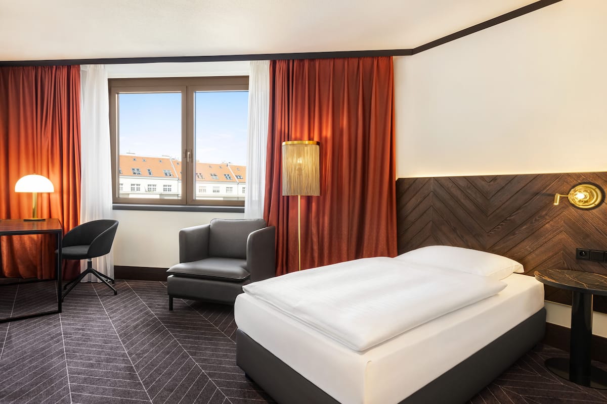 A hotel room with a bed, a desk, a chair, a lamp, and a view of buildings through a window.
