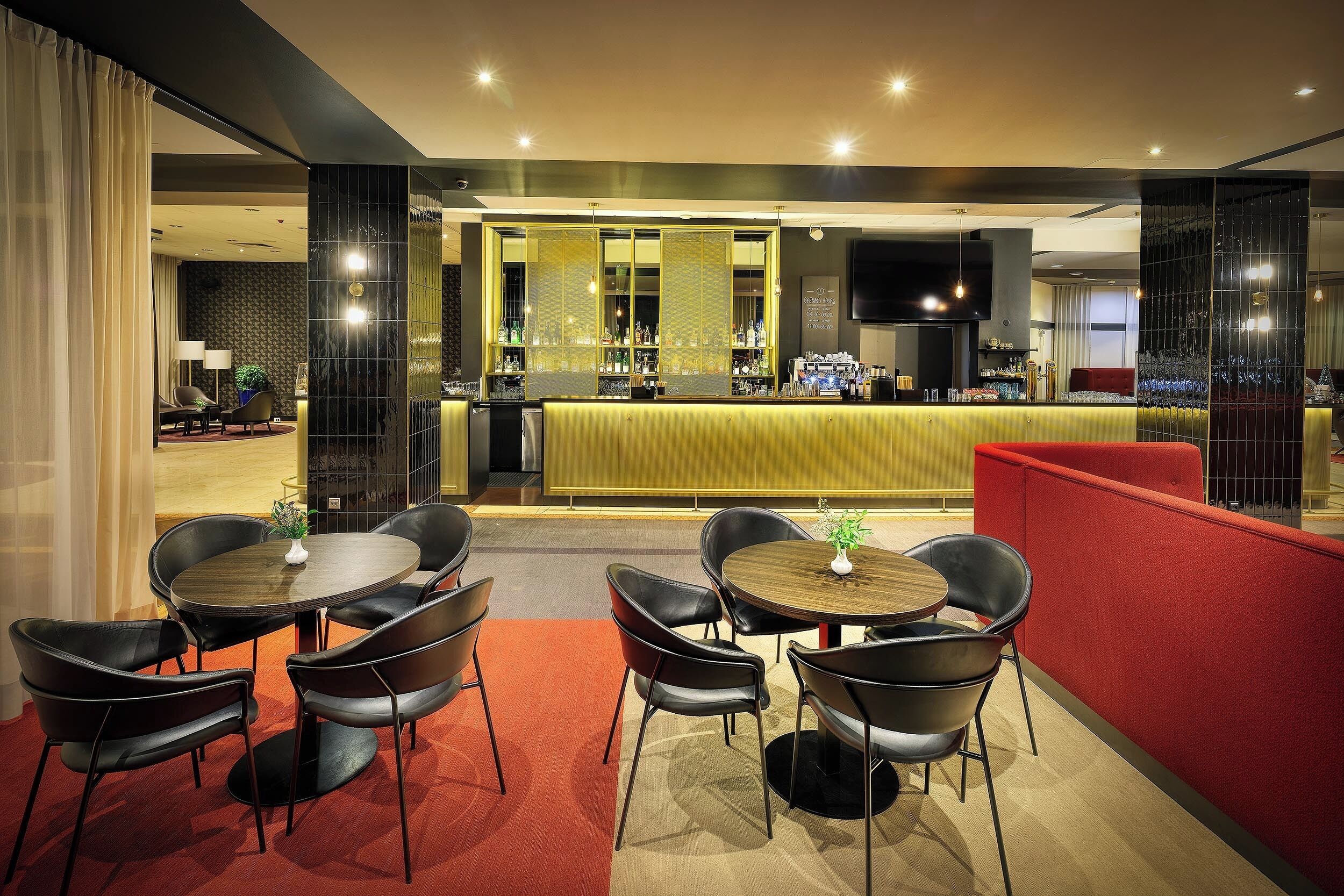 A modern bar area with a counter, bar stools, and a television screen. There are tables with chairs in the foreground, and a red partition separates the seating area from the bar.