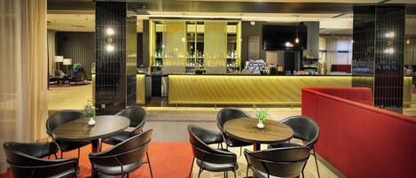 Un área de bar moderna con mostrador, taburetes y una pantalla de televisión. En primer plano hay mesas con sillas, y una separadora roja delimita la zona de estar del bar.