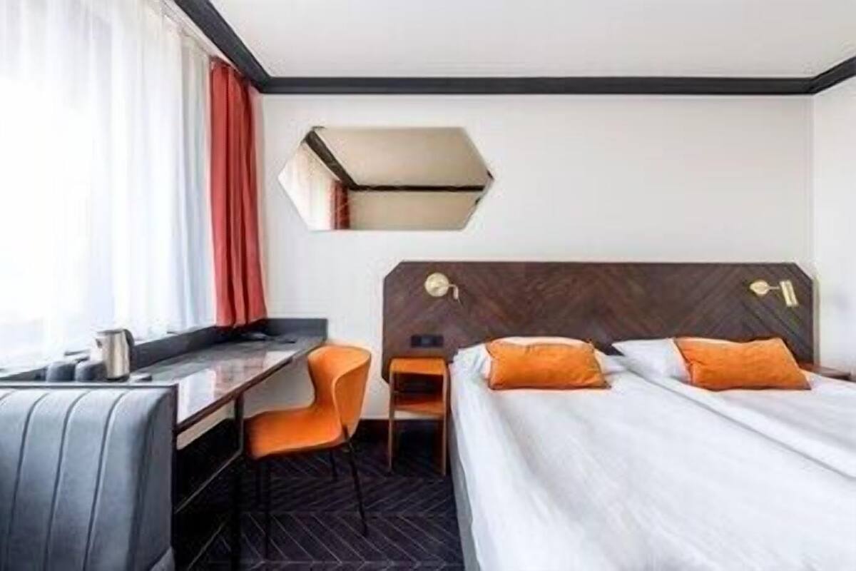 A hotel room with a bed, a desk, an orange chair, and a small table.
