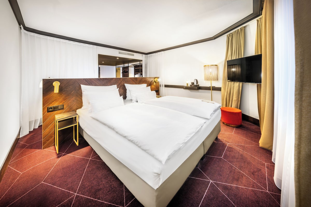 A hotel room with a large bed, a bedside table, a TV, and a red ottoman.